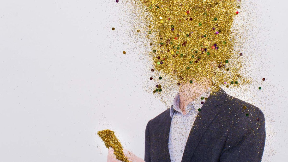 A man in a business suit looks at a gold phone with his head in an exploding cloud of gold dust.