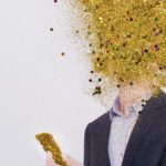 A man in a business suit looks at a gold phone with his head in an exploding cloud of gold dust.