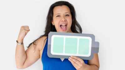 A smiling woman holds an arm in the air in triumph while also holding a graphic of a fully-charged battery in her other hand representing the Pilbara Minerals share price