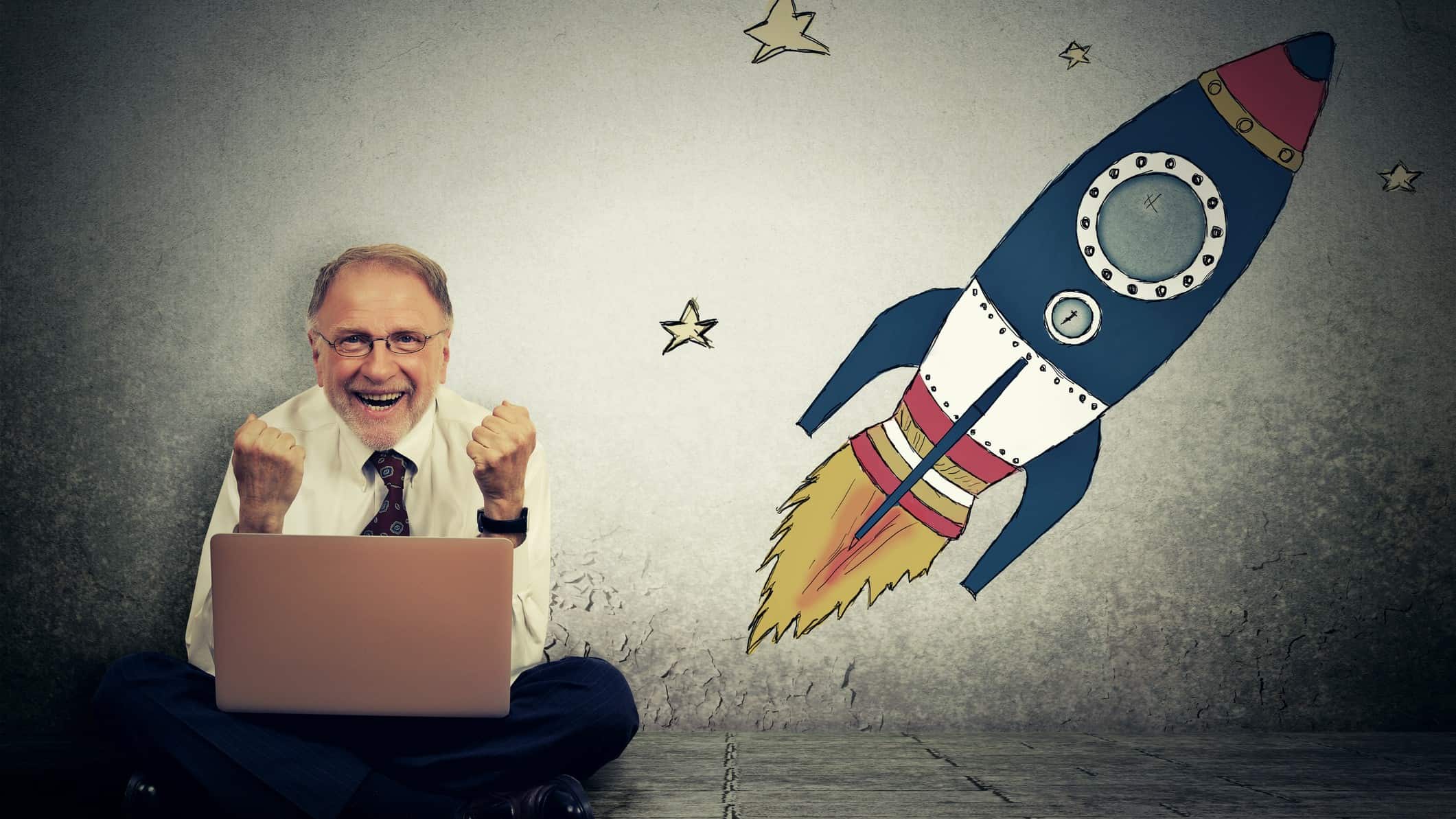 A male ASX investor sits cross-legged with a laptop computer in his lap with a slightly crazed, happy, excited look on his face while next to him a graphic of a rocket shoots upwards with graphics of stars scattered around it