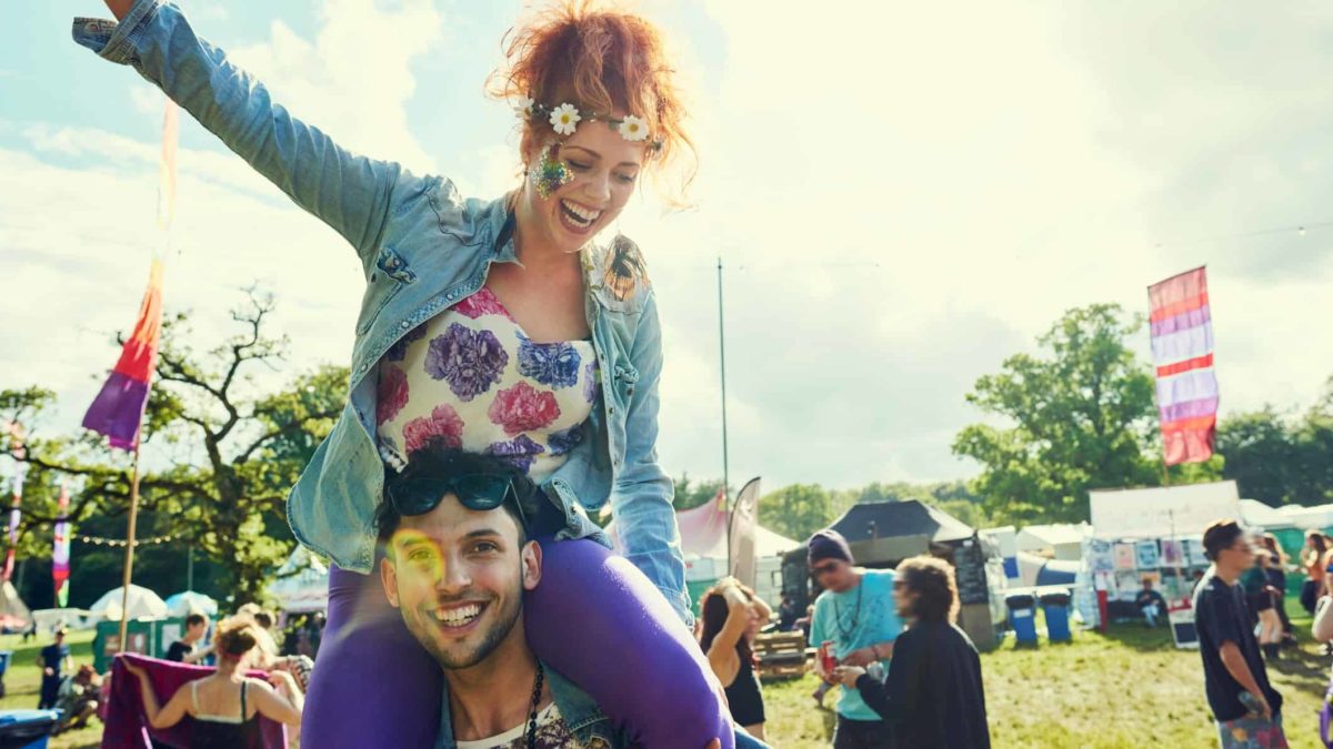 a woman wearing a flower garland sits atop the shoulders of a man celebrating a happy time in the outdoors with people talking in groups in the background, perhaps at an outdoor markets or music festival, in an image portraying young people enjoying freedom.