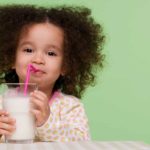 A cute young girl with curly hair sips a glass of milk through a straw with a smile on her face.