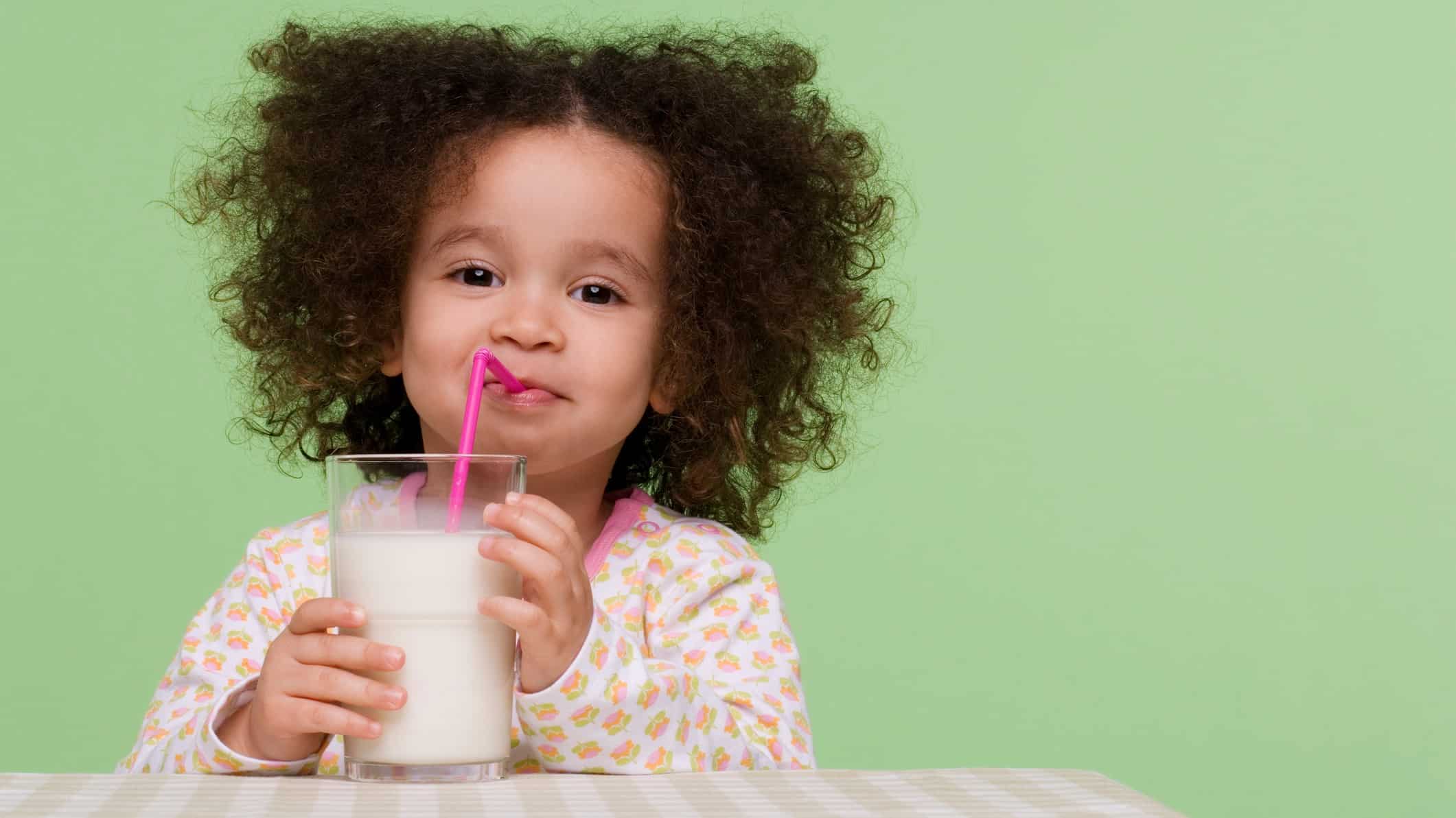 A cute young girl with curly hair sips a glass of milk through a straw with a smile on her face.