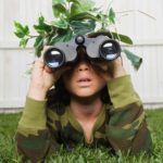 A child dressed in army clothes looks through his binoculars with leaves and branches on his head.