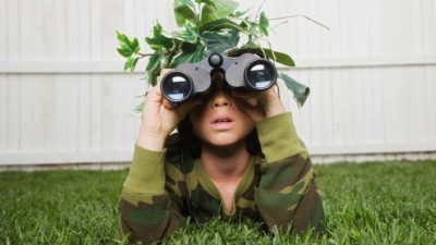 a child dressed in army fatigues lies on the ground in his backyard wearing leaves and branches on his head as camouflage and peering through a pair of binoculars in a soldier pose.
