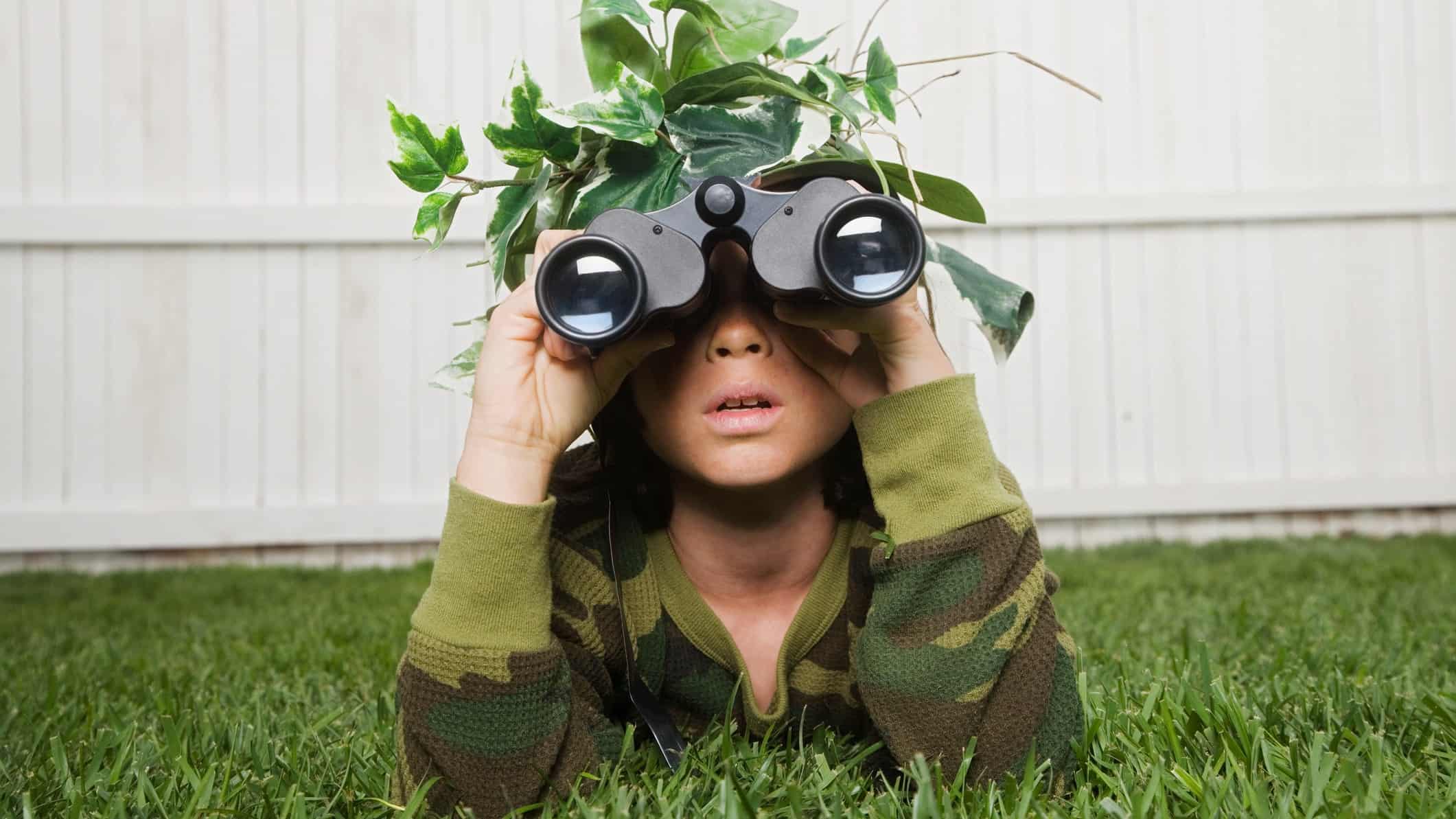 A child dressed in army clothes looks through his binoculars with leaves and branches on his head.