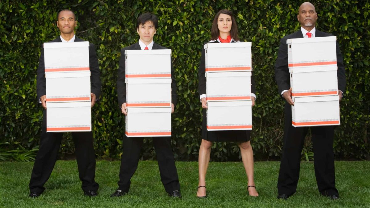 a group of four people wearing corporate uniforms stand in a line caring stacked boxes with unhappy looks on their faces.