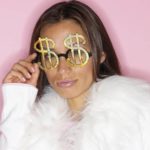 A woman in a fur coat adjusts her glasses made of gold dollar signs and pouts at the camera.