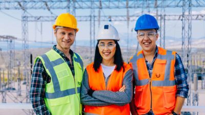 a group of three electricity workers stand smiling wearing hard hats and high visibility vests in front of an array of high voltage power equipment.