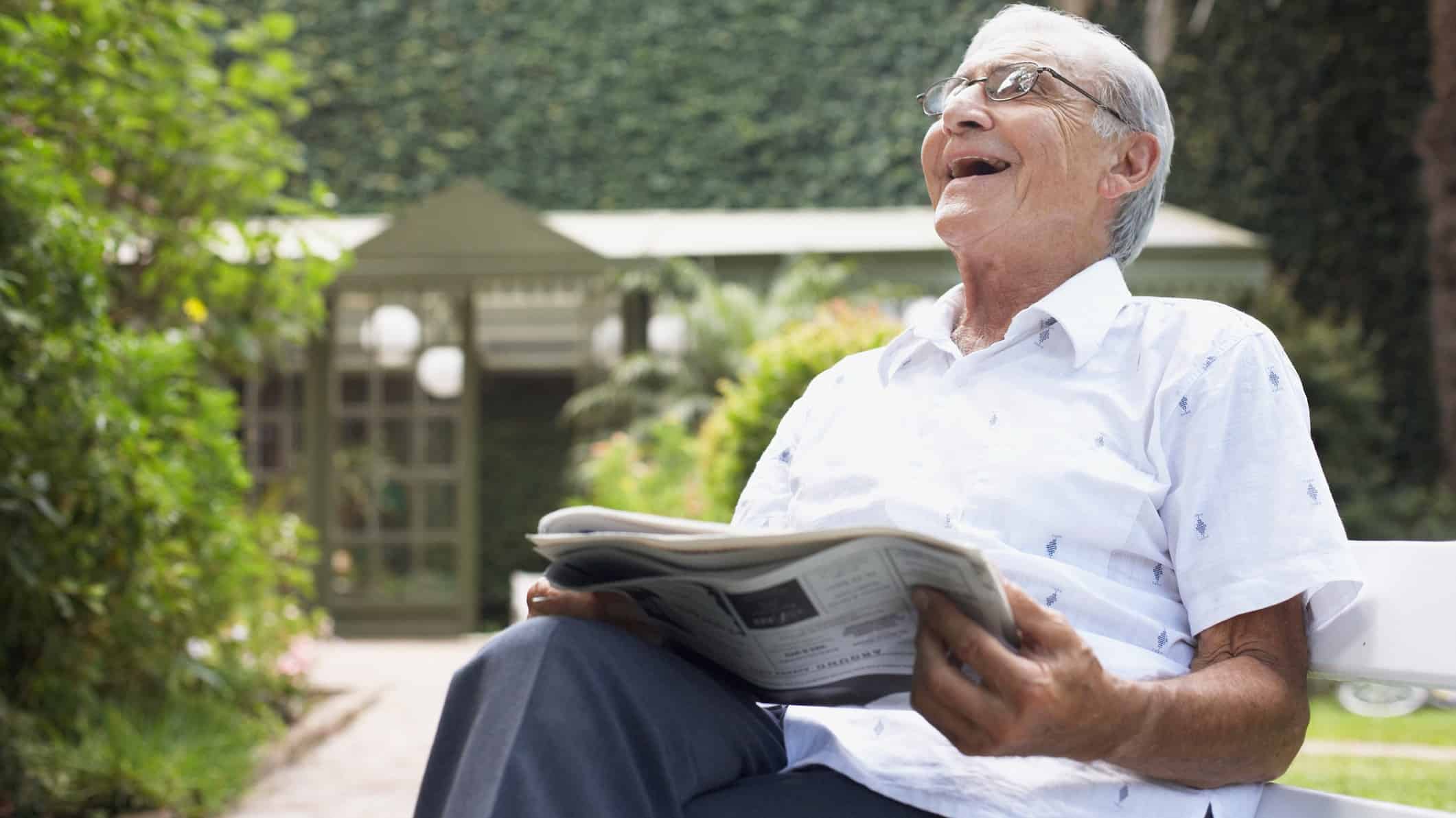 a happy investor, in this case an older gentleman, throws his head back and laughs while reading the newspaper in his garden.