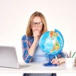 A woman with an open laptop holding a globe on a desk ponders something.