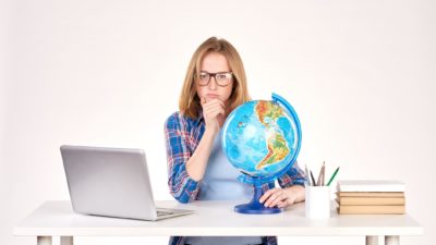 a woman sits at her desk looking puzzled and disappointed with her hand to her chin while an open laptop computer sits on one side of her and her hand is around the base of a globe of the world on the other side of her.