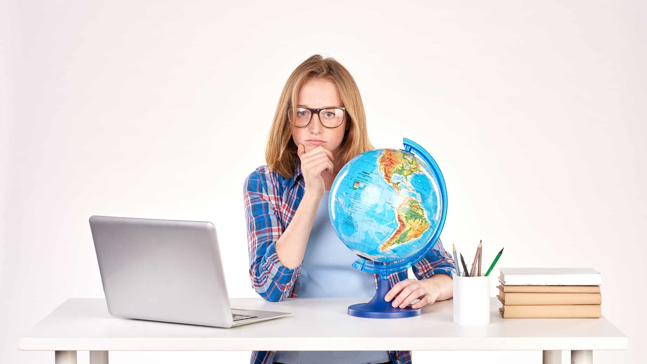 A woman with an open laptop holding a globe on a desk ponders something.