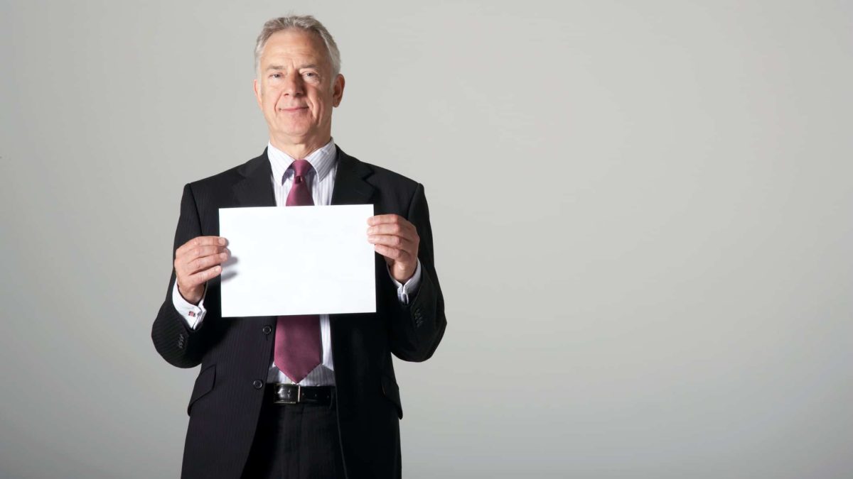 a man in a business suit holds a piece of paper in front of him as if revealing information.