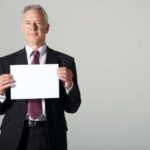 A man in a business suit holds a piece of paper in front of him as if revealing information.