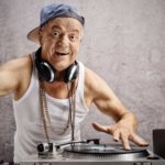 an older man dressed in singlet wearing thick neck chains and a side turned cap holds up two fingers while operating DJ mixing equipment with a record player and headphones around his neck.