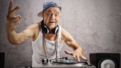an older man dressed in singlet wearing thick neck chains and a side turned cap holds up two fingers while operating DJ mixing equipment with a record player and headphones around his neck.
