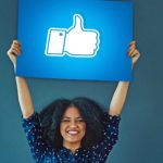 a woman holds a facebook like thumbs up sign high above her head. She has a very happy smile on her face.