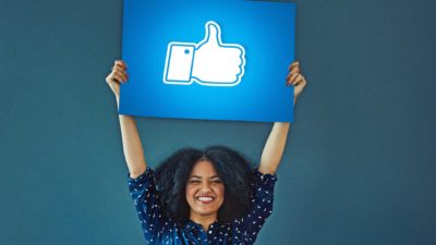 a woman holds a facebook like thumbs up sign high above her head. She has a very happy smile on her face.
