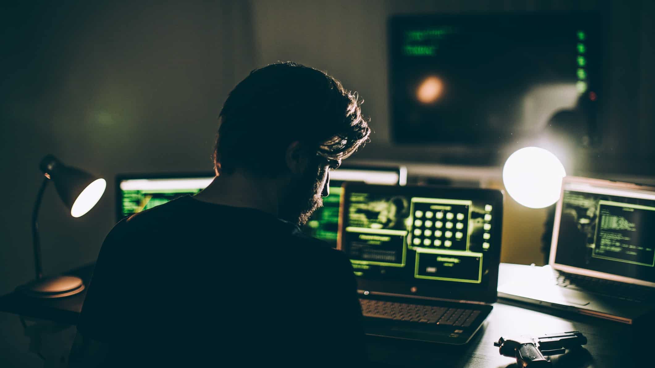 the back profile view of a man in a darkened room sitting in front of multiple computer screens, implying illegal hacking activity.