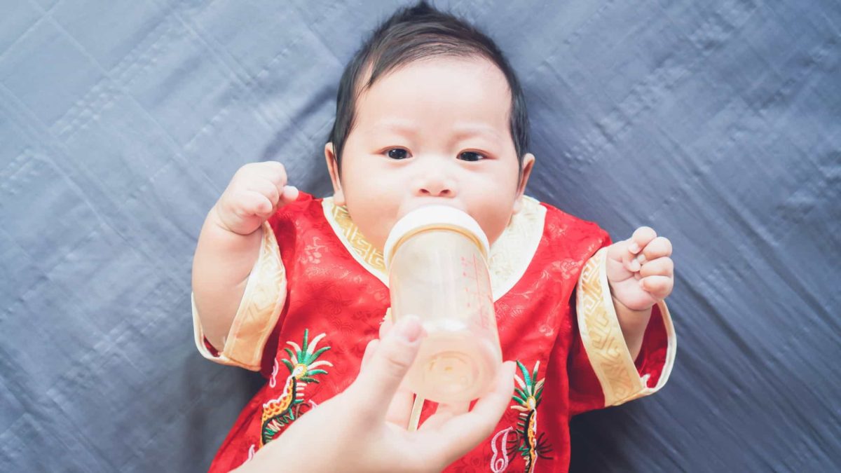 a cute small baby wearing a chinese embroidered outfit looks intently with hands outstretched as a hand holds a bottle of infant formula to his mouth.