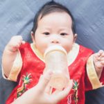 a cute small baby wearing a chinese embroidered outfit looks intently with hands outstretched as a hand holds a bottle of infant formula to his mouth.