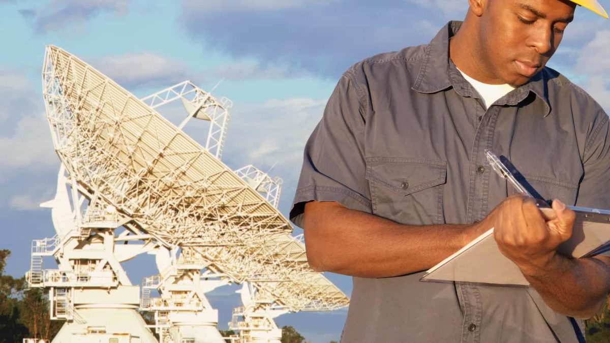 a worker with overalls and hard hat records information on a document with a large satellite dish in the background.