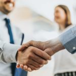 A close-up of a handshake depicting a business deal with one of the people in the background of the shot alongside a colleague looking pleased at the deal.