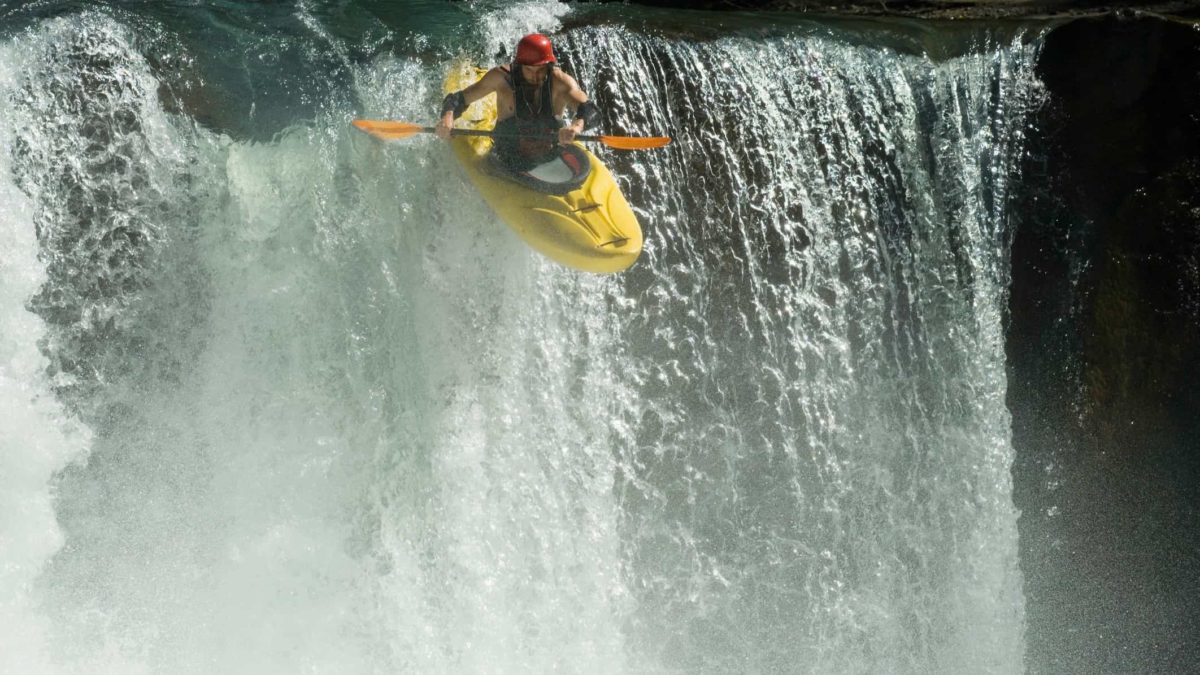 a white water rafter comes off the top of a large waterfall inevitably to hit the drop below.