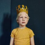 A boy with a gold crown stands stoically looking straight ahead.