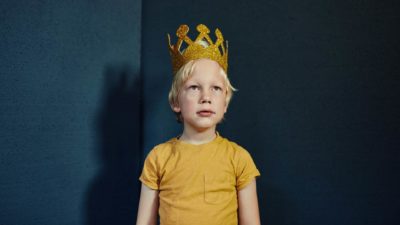 A boy with a gold crown stands stoically looking straight ahead.