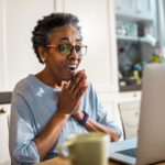 An older woman clasps her hands with joy, smiling at the news on her computer as she sits at her kitchen bench..