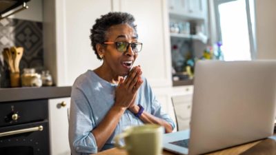 An older woman clasps her hands with joy, smiling at the news on her computer as she sits at her kitchen bench..