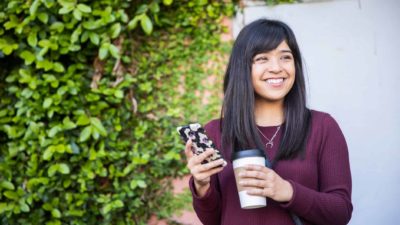 A woman smiles holding her phone and a takeaway coffee in front of a wall of green ivy.