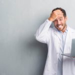 A man in a white coat holds a laptop in one hand and his head in the other, it's bad news.