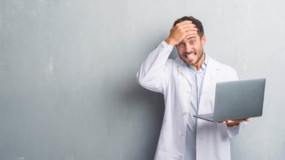 A man in a white coat holds a laptop in one hand and his head in the other, it's bad news.