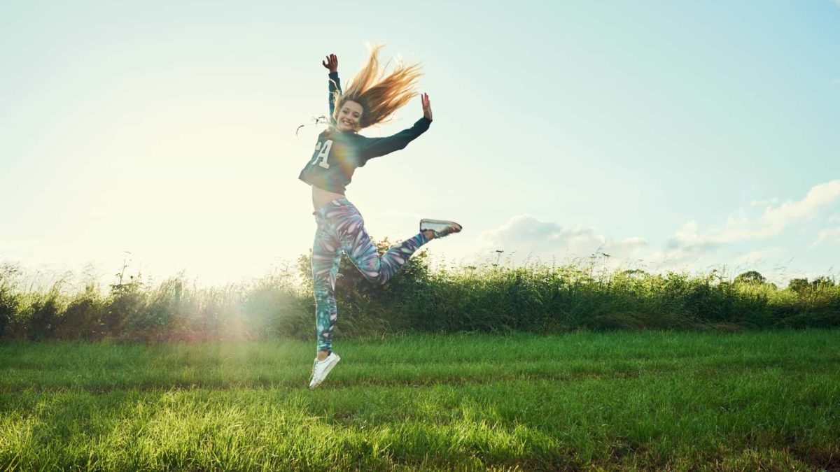 A woman leaps into the air with loads of energy, in a lush green field.