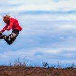A woman wearing a red jumper leaps into the air with sky behind her and earth beneath her.