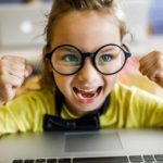 Little girl with big glasses on a laptop with a big smile on her face.