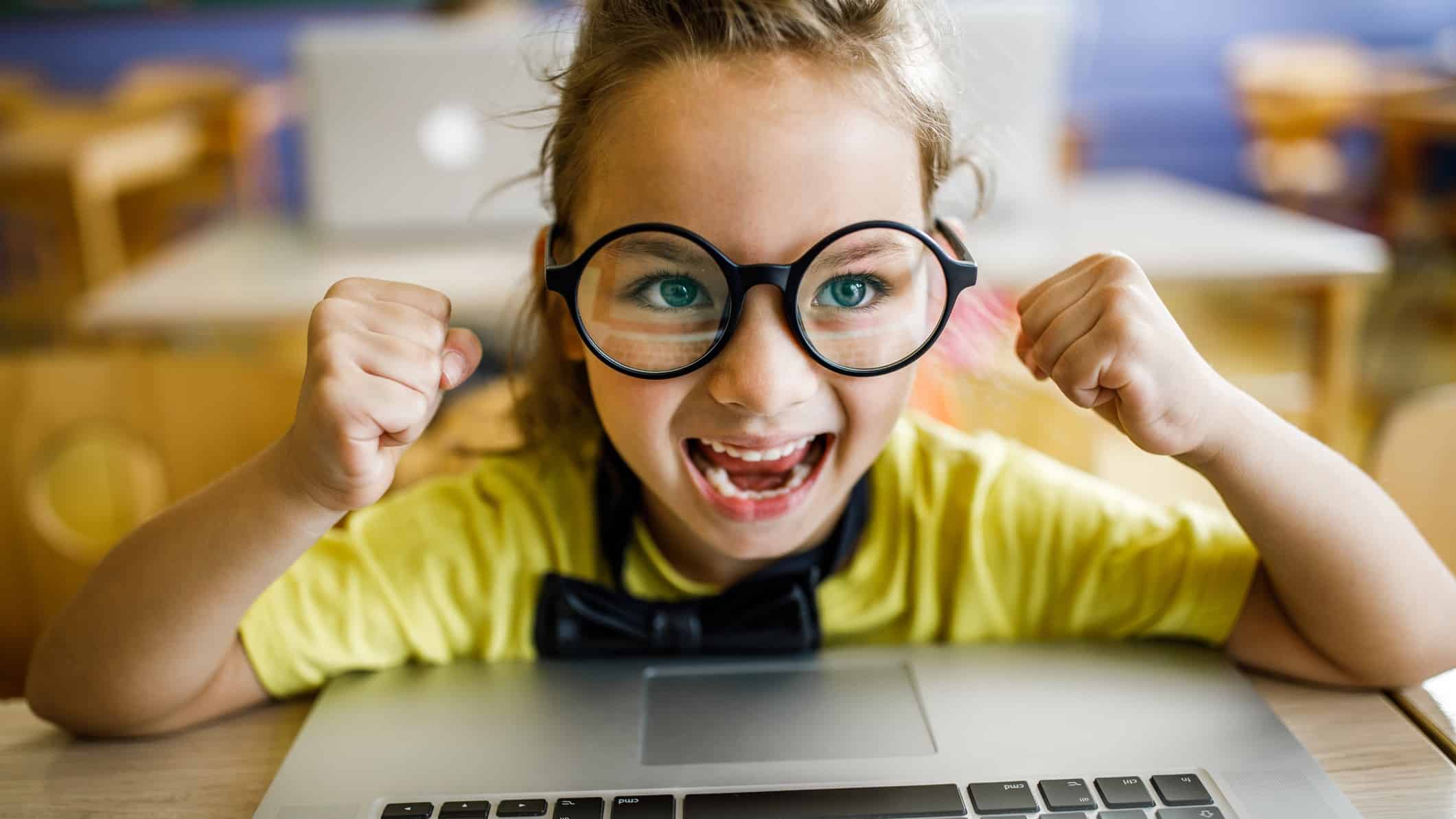 Little girl with big glasses on a laptop with a big smile on her face.