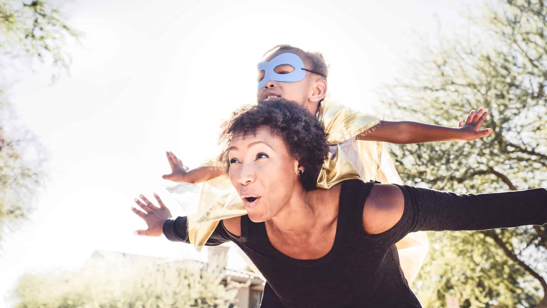 A mum lifts her superhero-face-mask-clad little girl on her shoulders as they both outstretch their arms in flight.