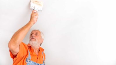An older man in an orange shirt paints the ceiling of a house.
