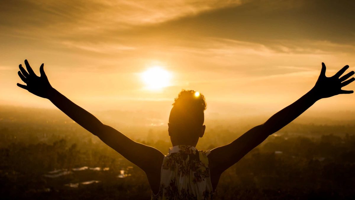 A woman stands triumphant with arms outstretched as she overlooks a city at sunset.