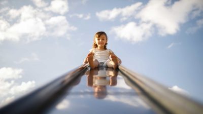 A little girl is about to launch down the slide with a blue sky and white clouds in the sky behind her.
