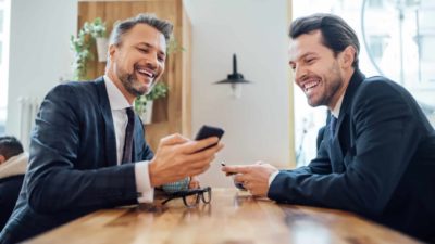 Two laughing male executives wearing dark suits chat across a timber lunch room table while one of them holds up his phone to show information.