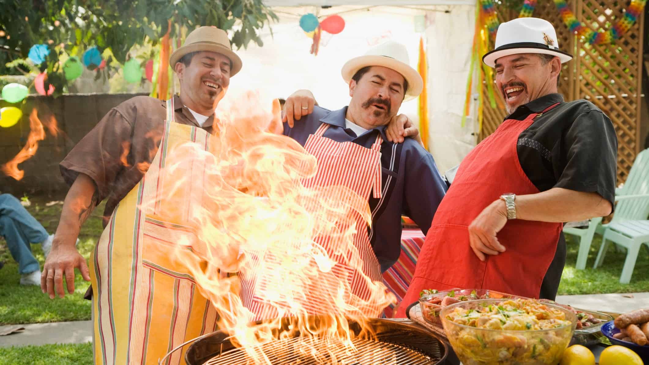 Three happy men with moustaches cooking on a BBQ with flames leaping up.