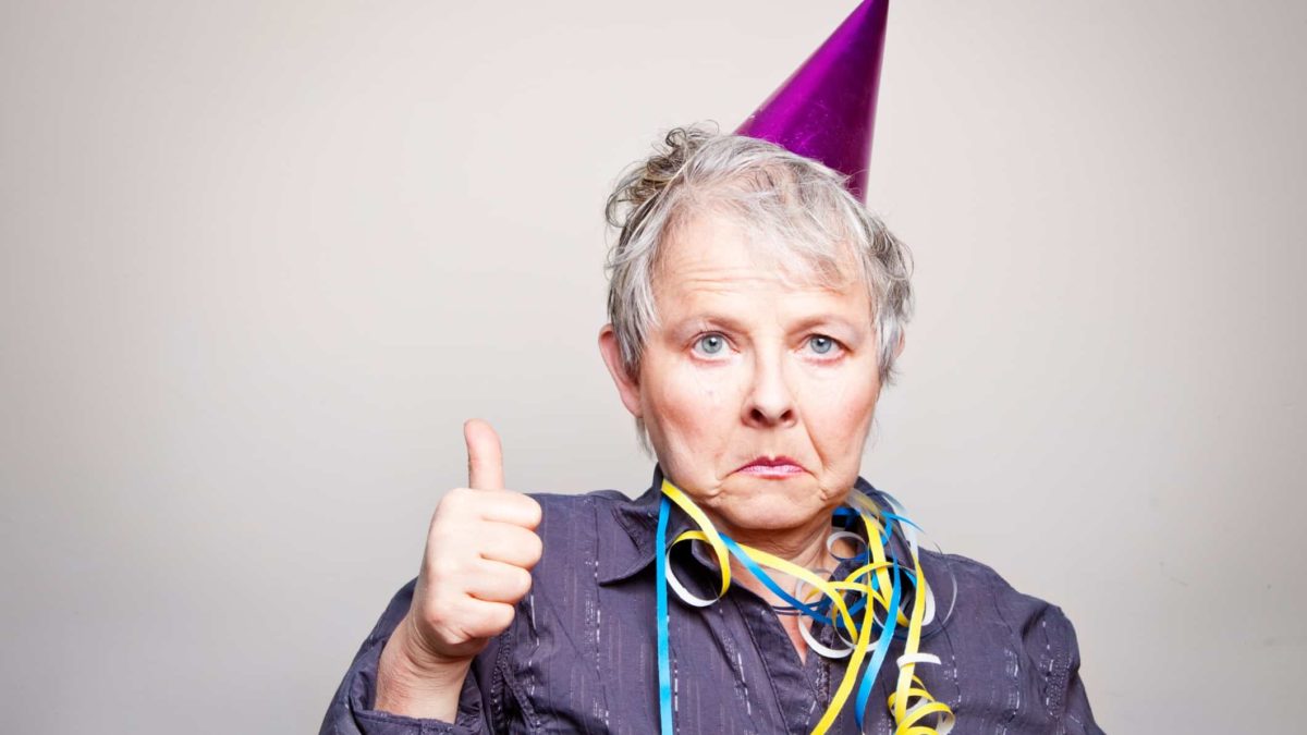An older woman wearing a party hat is giving a thumbs up, but she's not happy about it.
