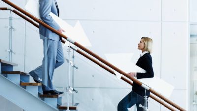 Two businesspeople walk in opposite directions on a staircase with arrows under their arms, one pointing up and one pointing down.