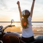 A woman sits on her motorbike looking out at the ocean with both fists in the air.