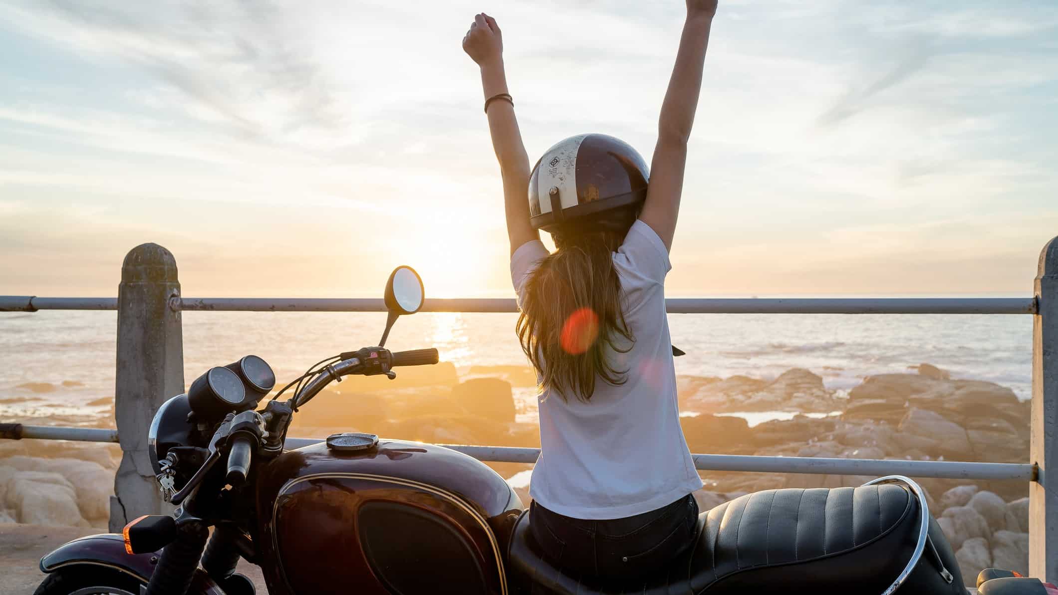 A woman sits on her motorbike looking out at the ocean with both fists in the air.